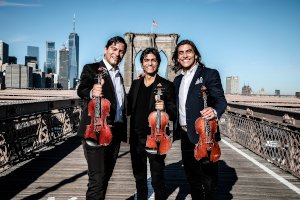 The three principal members of the Villalobos Brothers, standing on a pier with a city skyline in the background.
